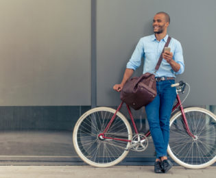 photo of main in jeans and blue shirt carrying a duffle bag and smart phone while smiling and leaning against a red city style bicycel