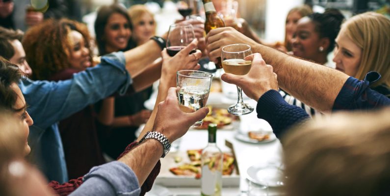 photo of dinner party and long table filled with table clinking glasses and bottles together while smiling