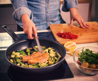 photo of man with tongs in kitchen cooking salmon filets with asparagus and zuchini in a pan with other chopped vegetables in bowl and cutting board next to stovetop